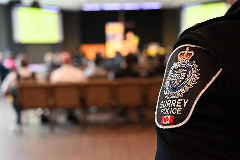 A close-up of a Surrey Police uniform sleeve showcases a badge, while a blurred audience engages in a presentation, highlighting community policing initiatives and collaboration.