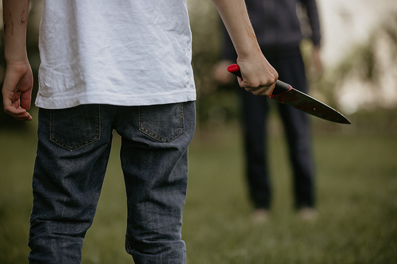 An individual stands with a bloody knife, facing another person in a tense outdoor setting. The image suggests conflict and the potential for violence, relevant to themes of policing and safety.